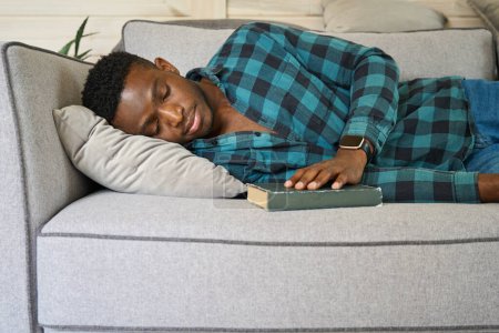 Foto de El hombre de pelo rizado se quedó dormido con un libro en el sofá, con una camisa a cuadros y vaqueros - Imagen libre de derechos
