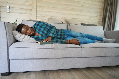 Photo for African American man dozed off with a book on the sofa, wearing a plaid shirt and jeans - Royalty Free Image