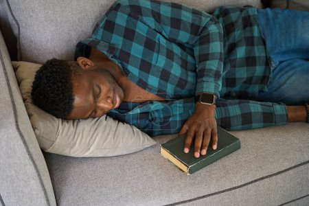 Photo for African American guy dozed off with a book on the sofa, wearing a plaid shirt and jeans - Royalty Free Image