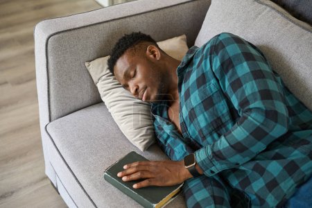 Téléchargez les photos : Jeune homme endormi avec un livre sur le canapé, portant une chemise à carreaux et un jean - en image libre de droit
