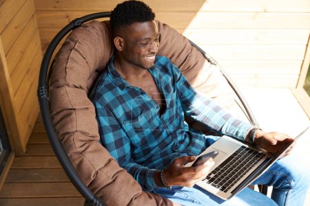 Foto de Hombre afroamericano se comunica en línea utilizando un ordenador portátil y teléfono móvil, él está sentado en una silla de jardín suave - Imagen libre de derechos