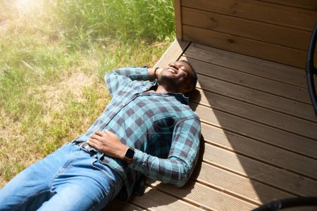 Téléchargez les photos : Joyeux mâle relaxant sur une terrasse en bois, il profite du matin d'été - en image libre de droit