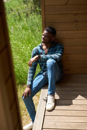 Foto de Chico con ropa cómoda está descansando en una terraza de madera, hay hierba verde exuberante en el césped - Imagen libre de derechos