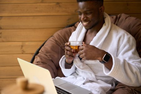 Foto de Sonriente hombre afroamericano se sienta con un ordenador portátil en una terraza de madera, té de la mañana se sirve en la mesa - Imagen libre de derechos