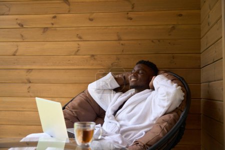 Téléchargez les photos : Un homme souriant s'assoit avec un ordinateur portable sur une terrasse en bois, le thé du matin est servi sur la table - en image libre de droit