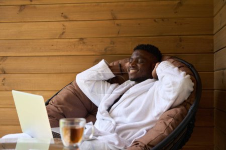 Foto de Chico alegre se sienta con un ordenador portátil en una terraza de madera, té de la mañana se sirve en la mesa - Imagen libre de derechos