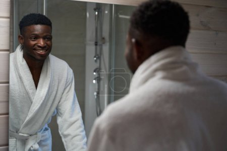 Photo for Man looks at himself in the mirror in the bathroom, he is in a cozy bathrobe - Royalty Free Image