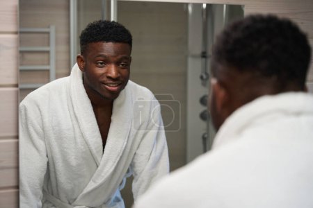 Photo for Male stands in front of the mirror in the bathroom, he is in a cozy bathrobe - Royalty Free Image