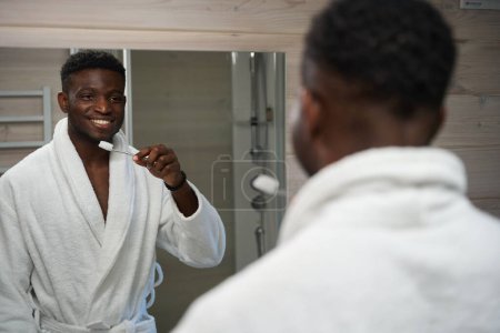 Téléchargez les photos : Homme se brosse les dents devant le miroir dans la salle de bain, il utilise une brosse à dents et du dentifrice - en image libre de droit