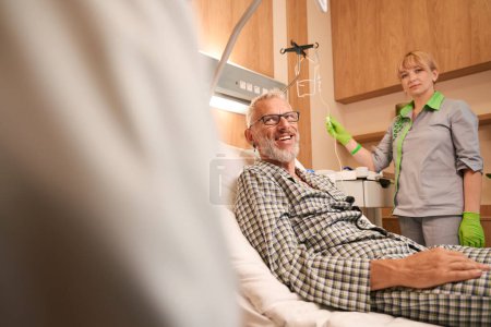 Téléchargez les photos : Le patient âgé communique avec le médecin dans une chambre d'hôpital, une infirmière prépare une intraveineuse pour lui à côté de lui - en image libre de droit