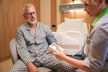 Téléchargez les photos : Femme médecin mesure la pression d'un homme âgé, ils sont situés dans une chambre d'hôpital confortable - en image libre de droit