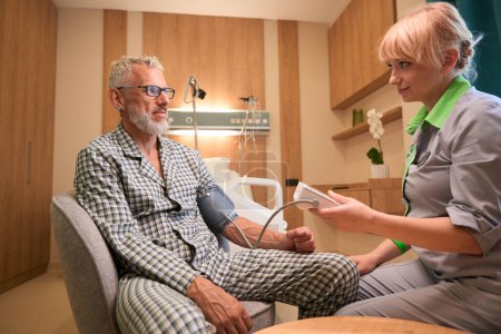 Téléchargez les photos : Infirmière blonde mesure la pression d'un vieil homme, ils sont situés dans une chambre d'hôpital confortable - en image libre de droit