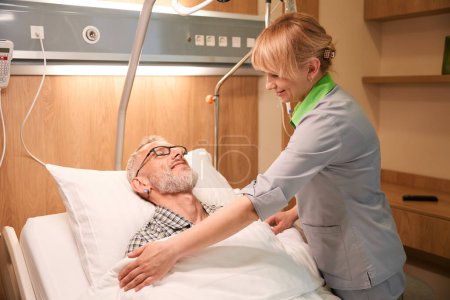 Téléchargez les photos : Jeune infirmière s'occupe d'un patient âgé, l'homme s'allonge sur un lit mobile spécial - en image libre de droit