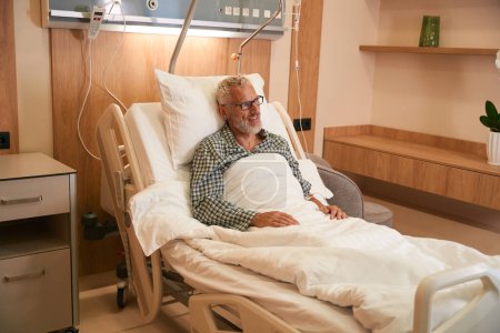 Téléchargez les photos : Patient âgé assis confortablement dans une chambre d'hôpital, il y a un stand avec un compte-gouttes à côté du lit - en image libre de droit