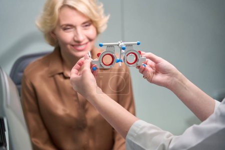 Téléchargez les photos : Ophtalmologiste met sur un dispositif spécial pour diagnostiquer les yeux d'une femme, une patiente dans un chemisier brun - en image libre de droit
