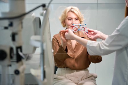 Téléchargez les photos : Une patiente en chemisier marron met un dispositif spécial pour vérifier la vision, à côté d'un jeune médecin - en image libre de droit