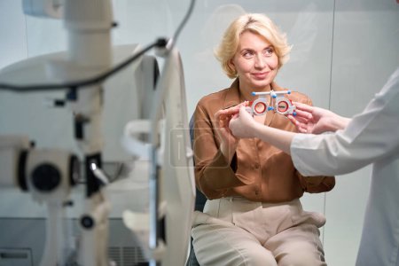 Téléchargez les photos : Une patiente en blouse brune met un dispositif spécial pour tester sa vision, à côté d'un jeune médecin - en image libre de droit