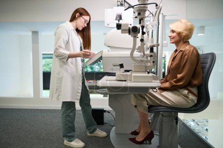 Foto de Médico pelirrojo está consultando a una señora de mediana edad en el centro de diagnóstico, hay dispositivos oftalmológicos modernos en la habitación - Imagen libre de derechos