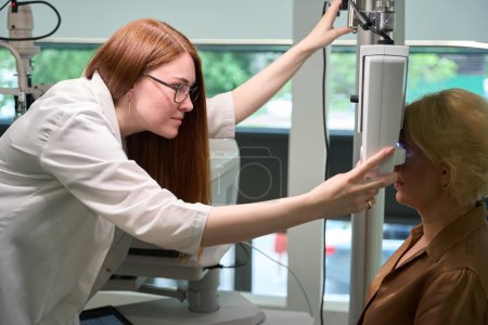 Téléchargez les photos : Médecin aux cheveux roux utilise un dioptrimètre dans son travail, le client est assis près de l'appareil - en image libre de droit