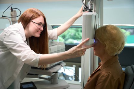 Photo for Pretty ophthalmologist uses a dioptermeter in her work, the client sits near the device - Royalty Free Image