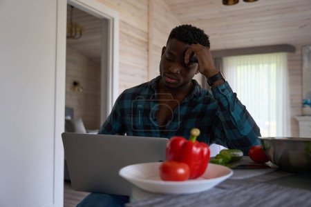 Téléchargez les photos : Homme aux cheveux bouclés assis avec un ordinateur portable dans la cuisine, il y a de la vaisselle et de la nourriture sur la table - en image libre de droit