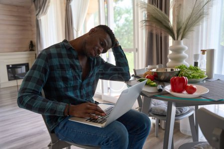Téléchargez les photos : Homme inquiet en vêtements décontractés se trouve avec un ordinateur portable dans la cuisine, il ya de la vaisselle et de la nourriture sur la table - en image libre de droit