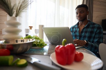 Foto de Joven se sienta con un ordenador portátil en la mesa de la cocina, hay verduras para ensalada en la mesa - Imagen libre de derechos