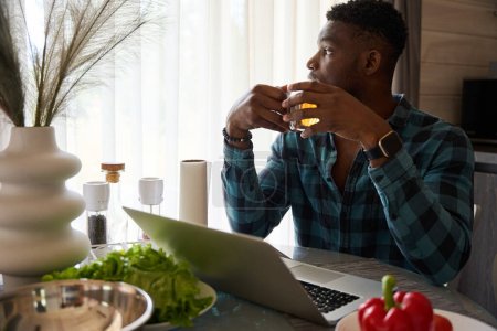 Foto de Hombre con taza de té en la mesa de la cocina, en la mesa hay un ordenador portátil y verduras para ensalada - Imagen libre de derechos
