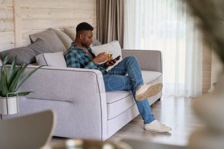 Photo for Guy in a checkered shirt with mobile phone is sitting on the sofa, he is located in a bright room - Royalty Free Image