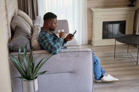Photo for African American guy with a mobile phone sits on the sofa, he is located in a bright room - Royalty Free Image