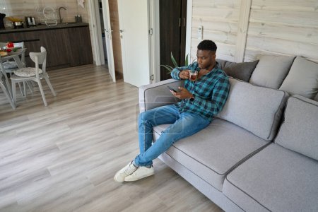 Photo for African American male with cup of tea and mobile phone sits on sofa, he is located in a bright room - Royalty Free Image