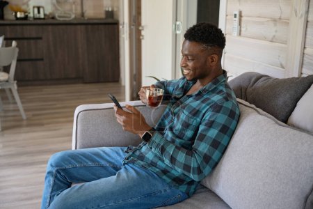 Téléchargez les photos : Jeune homme avec une tasse de thé et un téléphone portable sur le canapé, il est situé dans une pièce lumineuse - en image libre de droit