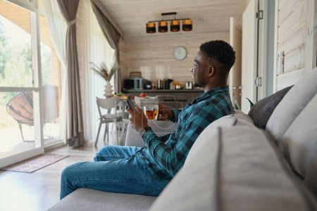 Photo for Young man is relaxing with cup of tea and mobile phone on sofa, he is located in a bright room - Royalty Free Image