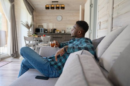 Photo for Young man is relaxing with a cup of tea on the sofa, he is located in a bright room - Royalty Free Image
