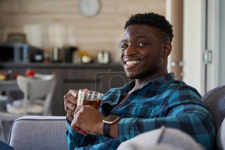 Foto de Hombre afroamericano con un reloj inteligente en la mano, se sentó con una taza de té en el sofá - Imagen libre de derechos