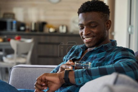 Photo for African American male looks at smart watch on his hand, he is sitting on the sofa with cup of tea - Royalty Free Image