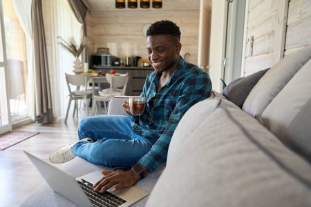 Photo for Smiling male sits with a cup of tea on a cozy sofa, he works online using a laptop - Royalty Free Image