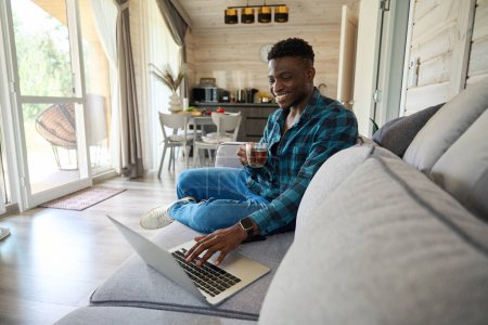 Téléchargez les photos : Jeune homme travaille en ligne à l'aide d'un ordinateur portable, il est assis avec une tasse de thé sur un canapé confortable - en image libre de droit