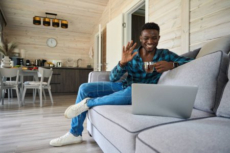Téléchargez les photos : Guy communique en ligne sur un ordinateur portable, il s'assoit avec une tasse de thé sur un canapé confortable - en image libre de droit