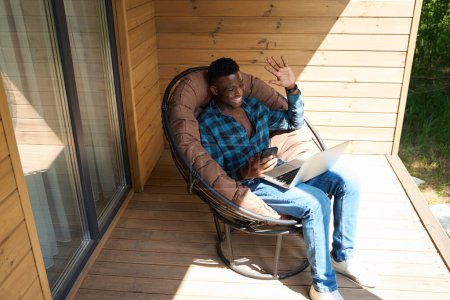 Photo for African American guy communicates online on a laptop, he is sitting in a soft garden chair - Royalty Free Image