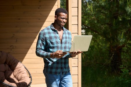 Foto de Hombre afroamericano salió a una terraza de madera con un ordenador portátil, llevaba ropa cómoda para el hogar - Imagen libre de derechos