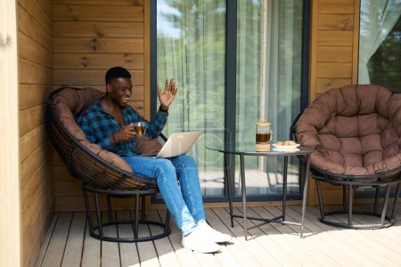 Photo for Nice guy in a checkered shirt sits with a laptop on a wooden terrace, tea is served on the table - Royalty Free Image