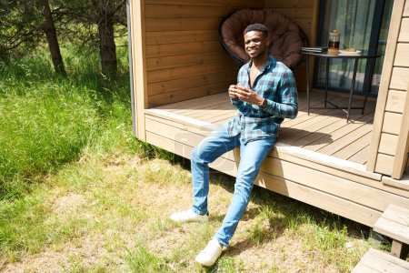 Photo for African American man on a wooden terrace with a cup of tea, he enjoys a beautiful morning - Royalty Free Image