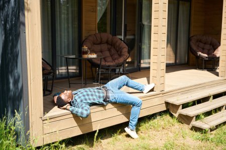 Photo for Male in home clothes relaxes on a wooden terrace, he enjoys the summer morning - Royalty Free Image