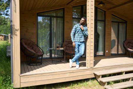 Téléchargez les photos : Afro-Américain debout sur la terrasse ensoleillée, thé du matin est servi sur la table - en image libre de droit