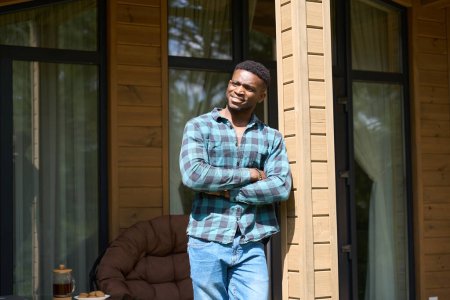 Téléchargez les photos : Afro-Américain se tient sur la terrasse ensoleillée, thé du matin est servi sur la table - en image libre de droit
