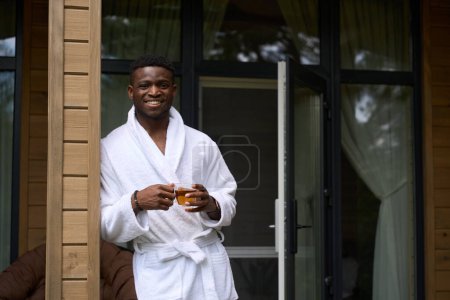 Foto de Chico afroamericano saluda a la mañana en una terraza de madera con una taza de té, él está en una acogedora túnica de rizo - Imagen libre de derechos