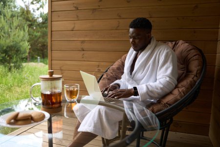 Foto de Chico afroamericano se sienta con el ordenador portátil en una silla en una terraza de madera, té de la mañana se sirve en la mesa - Imagen libre de derechos