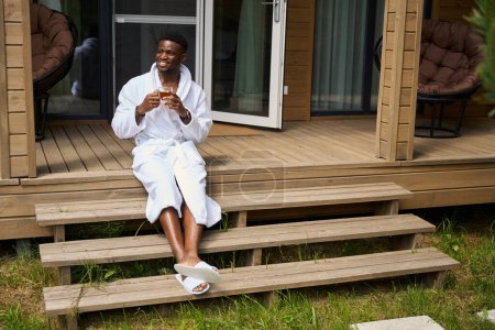 Foto de Sonriente chico afroamericano en el porche con té de la mañana, él está en una acogedora túnica de felpa - Imagen libre de derechos