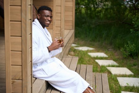 Foto de Feliz chico afroamericano en el porche con té de la mañana, él está en una acogedora túnica de felpa - Imagen libre de derechos